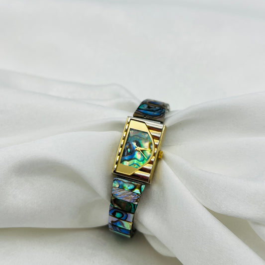 A gold-toned watch with an abalone shell band, displayed on a white fabric surface.